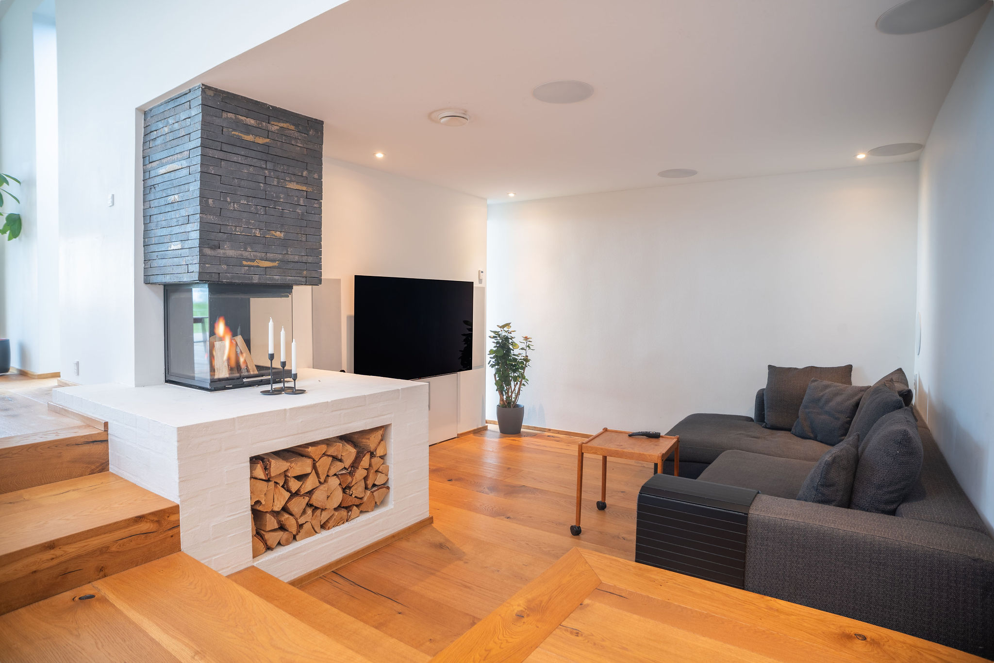 Moderne stue med trægulve, en mørkegrå sektionssofa, et fladskærms-tv og stilfulde Airbnb-detaljer - en potteplante og en dobbeltsidet pejs med stablet brænde under og stearinlys ovenpå - perfekt til enhver fotografs portefølje.