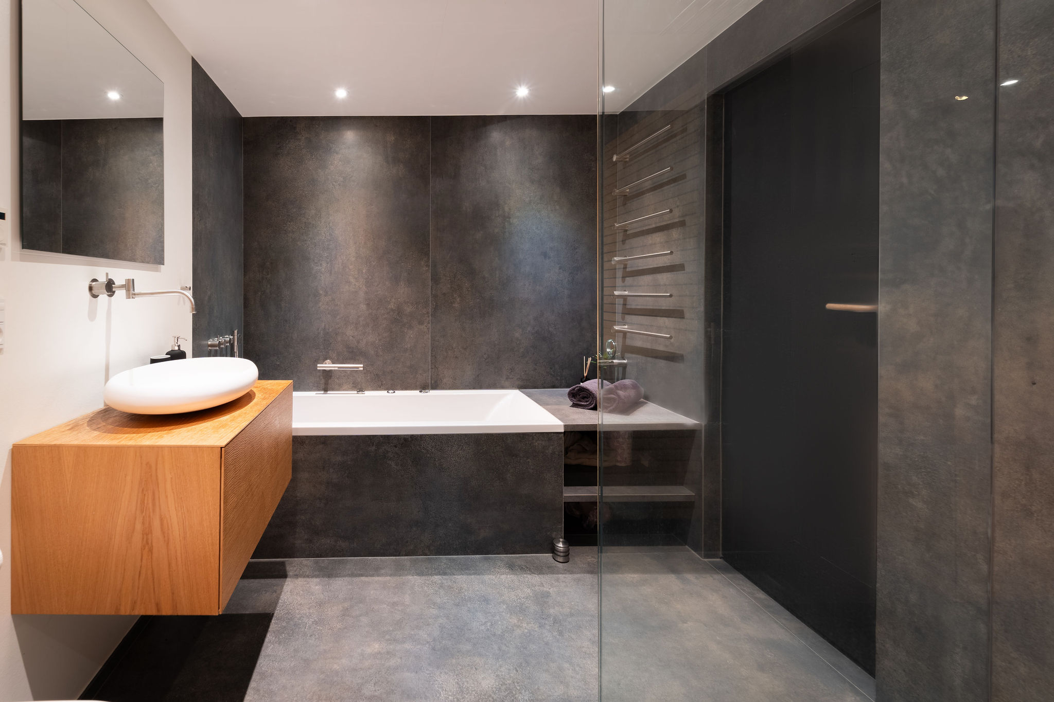 Moderne Airbnb-badeværelse med minimalistisk design, med en flydende trævask, en rund hvid vask, et badekar med mørke teksturerede vægge - perfekt til enhver fotograf, der søger stil. Det veloplyste rum har en glasvæg og en opvarmet håndklædetørrer.