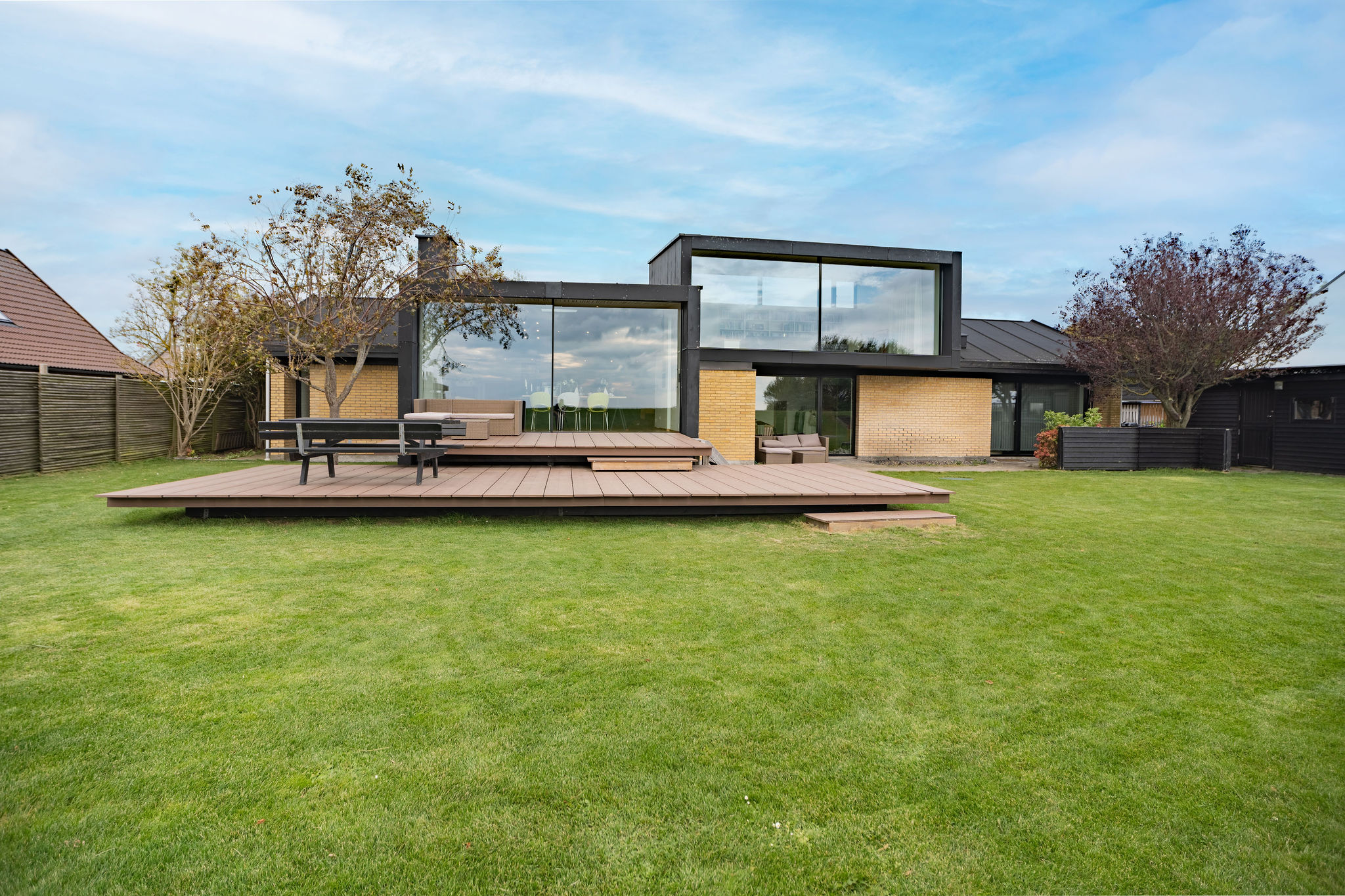 Moderne hus med store glasvinduer og en rummelig træterrasse i en græsklædt baghave - perfekt til dit næste Airbnb-ophold. Billedet er taget af en dygtig fotograf og har frodige træer og en blå himmel i baggrunden.