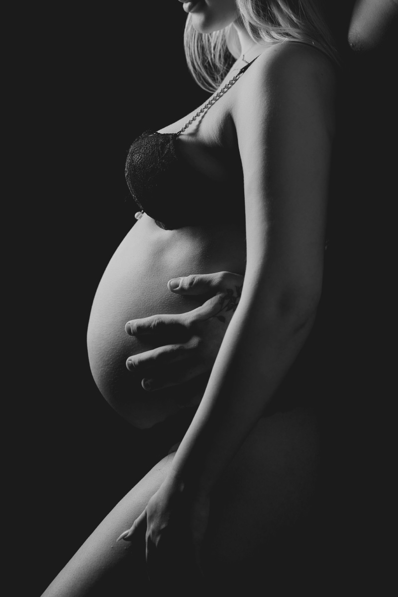 En gravid kvinde i sort lingeri bliver blidt omfavnet bagfra af en partner, hvis hænder hviler på hendes bare mave. Denne gravid fotosession i studie er blødt belyst, hvilket fremhæver kurverne og skaber en varm, intim atmosfære.