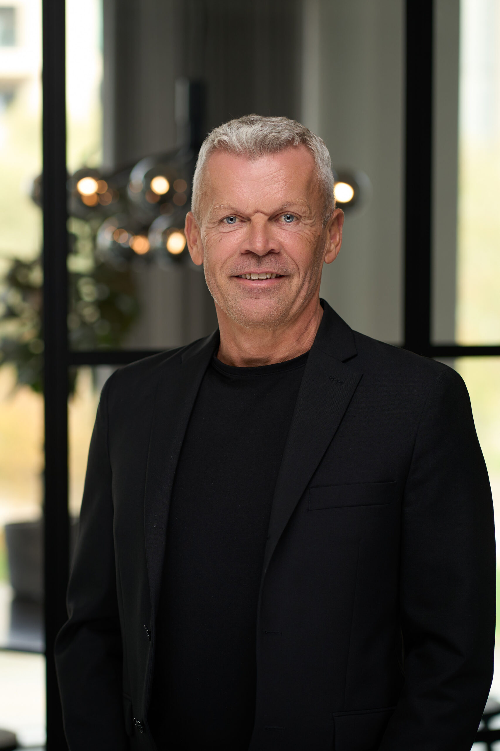 Professionelt portræt af en mand iført en sort blazer og sort t-shirt, taget i et moderne kontormiljø med slørede detaljer af glasvægge og belysning i baggrunden.