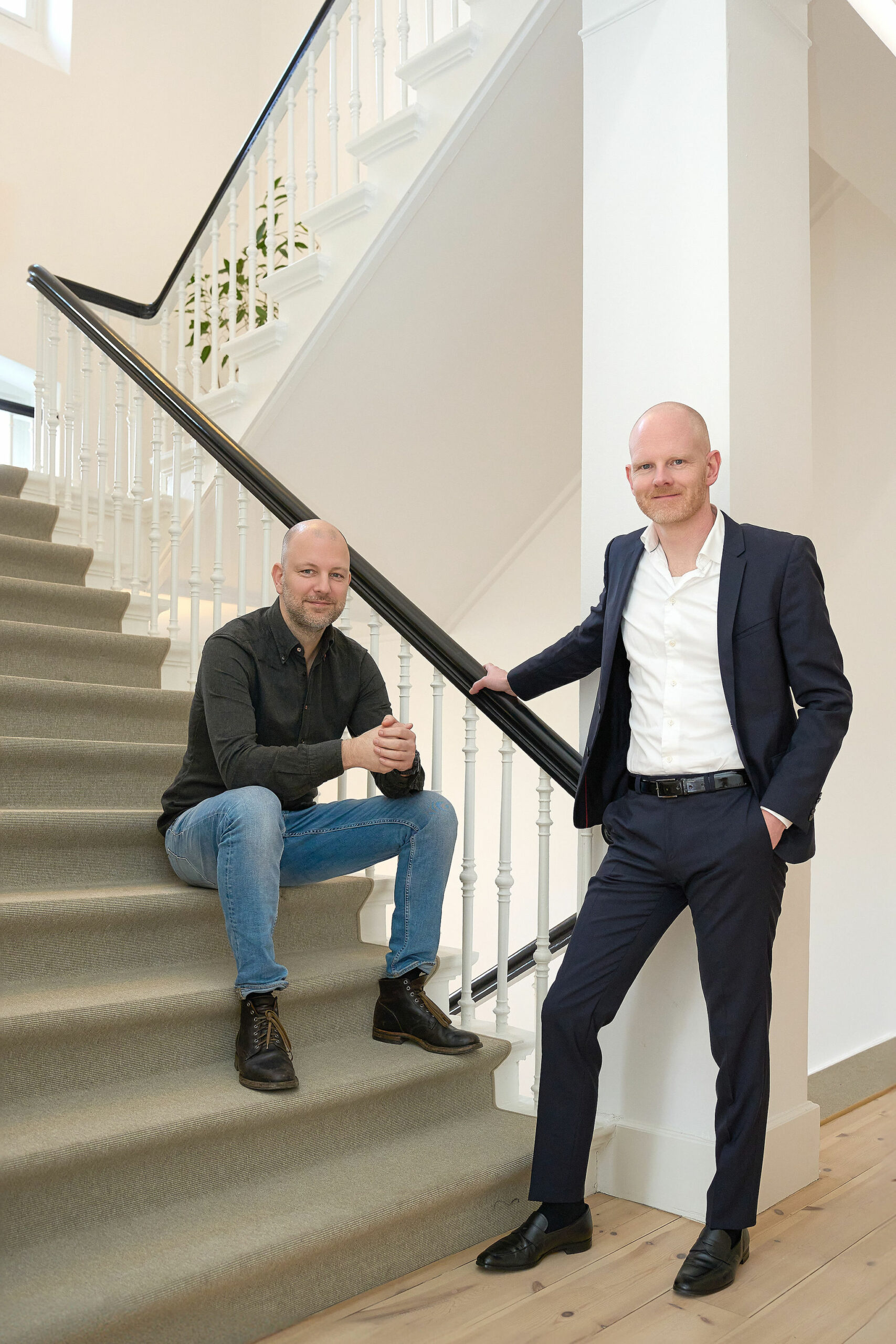 To mænd med barberede hoveder poserer ved en trappe; den ene sidder afslappet på trinene i en mørk skjorte og jeans, mens den anden står ved gelænderet i et marineblåt jakkesæt og en hvid skjorte og smiler selvsikkert til et moderne forretningsportræt.
