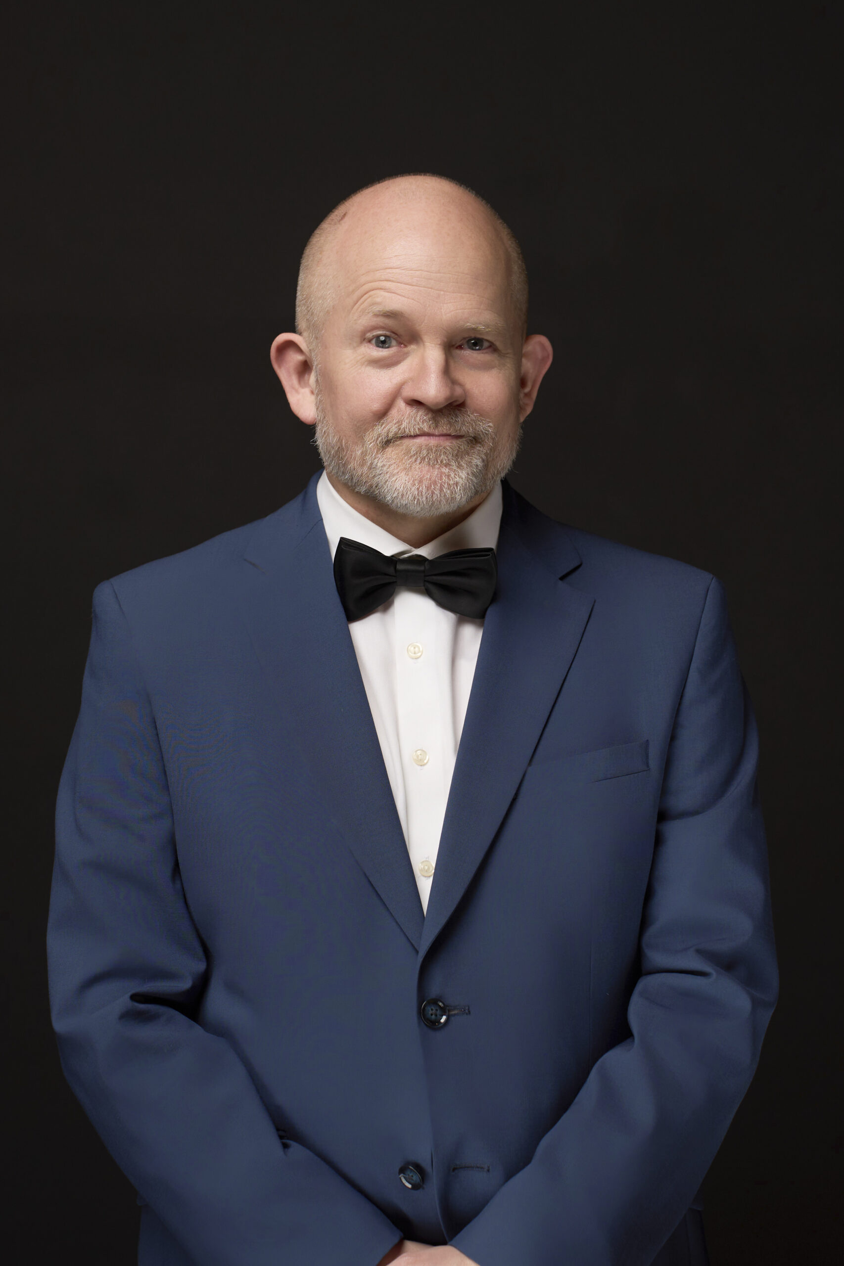 Portræt af skuespiller: En midaldrende mand med skaldet hoved og gråt skæg står på en sort baggrund, klædt i blåt jakkesæt og butterfly, med hænderne løst foldet, mens han smiler lidt. fotograf: Weigang Fotografi.