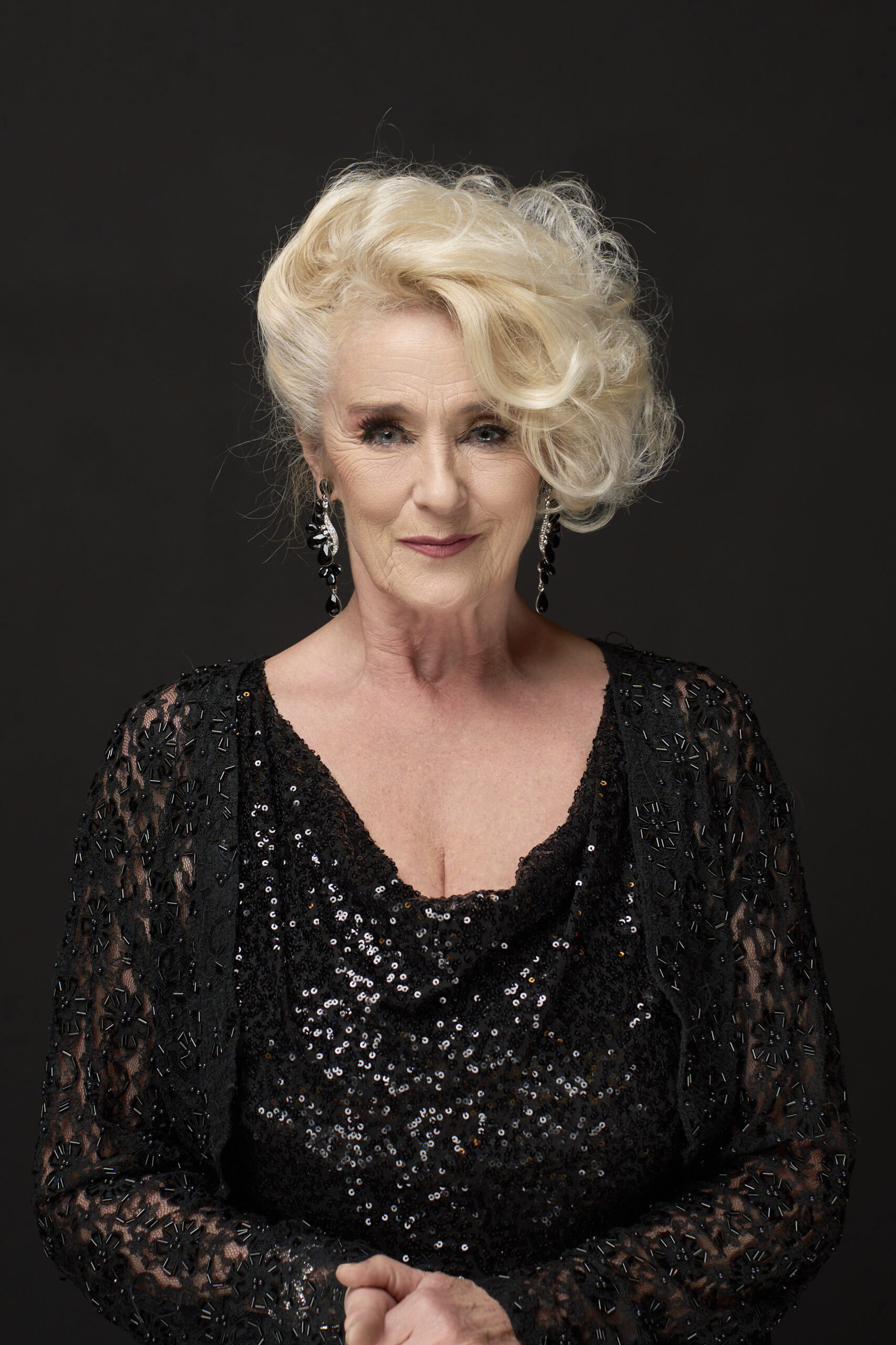 Portræt af skuespiller: En ældre kvinde med kort, krøllet blondt hår, iført en glitrende sort top og hængende øreringe, står foran en mørk baggrund og smiler blidt til kameraet. Fotograf: Weigang Fotografi.