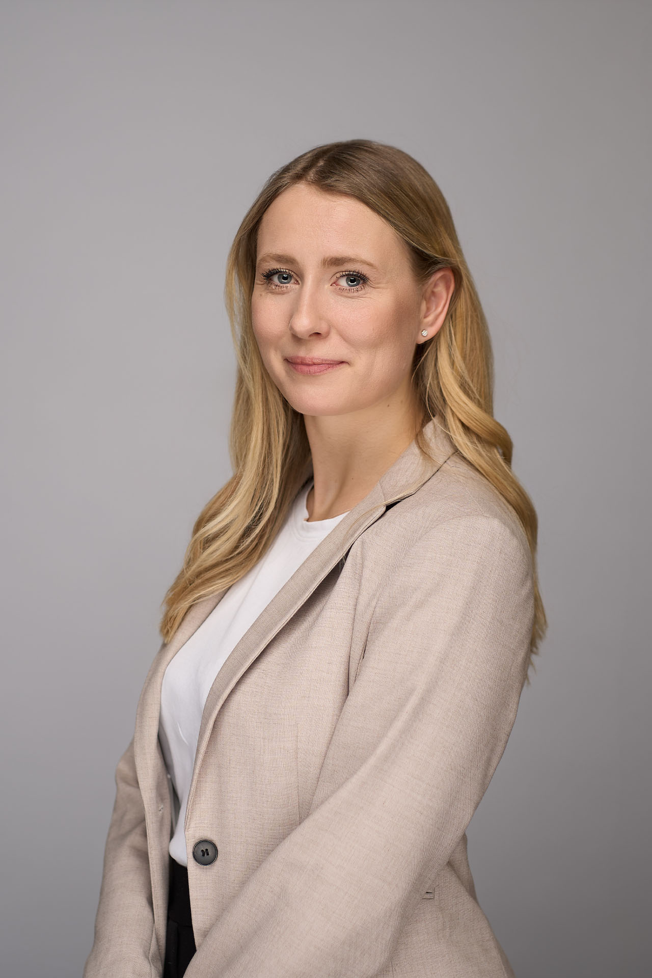 En kvinde med langt blondt hår er iført en lys beige blazer over en hvid top. Hun står foran en almindelig lysegrå baggrund og smiler blidt til kameraet i dette elegante portræt.