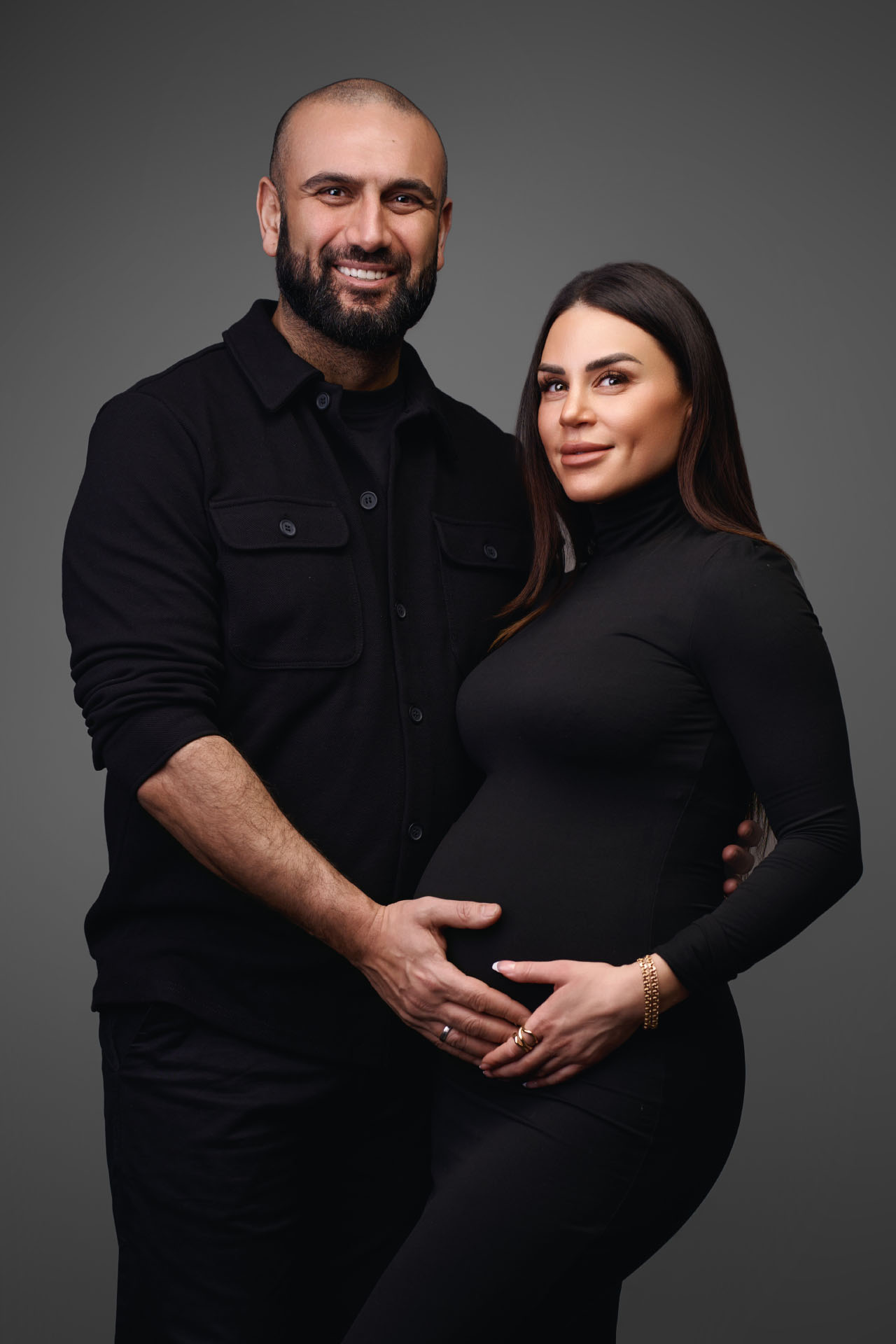 Et smilende par poserer sammen i en gravid fotosession, begge klædt i sort. Manden hviler blidt sine hænder på kvindens gravide mave, mens hun smiler blidt og holder hans hænder. Baggrunden er enkel og mørk og skaber et klassisk studie-look.