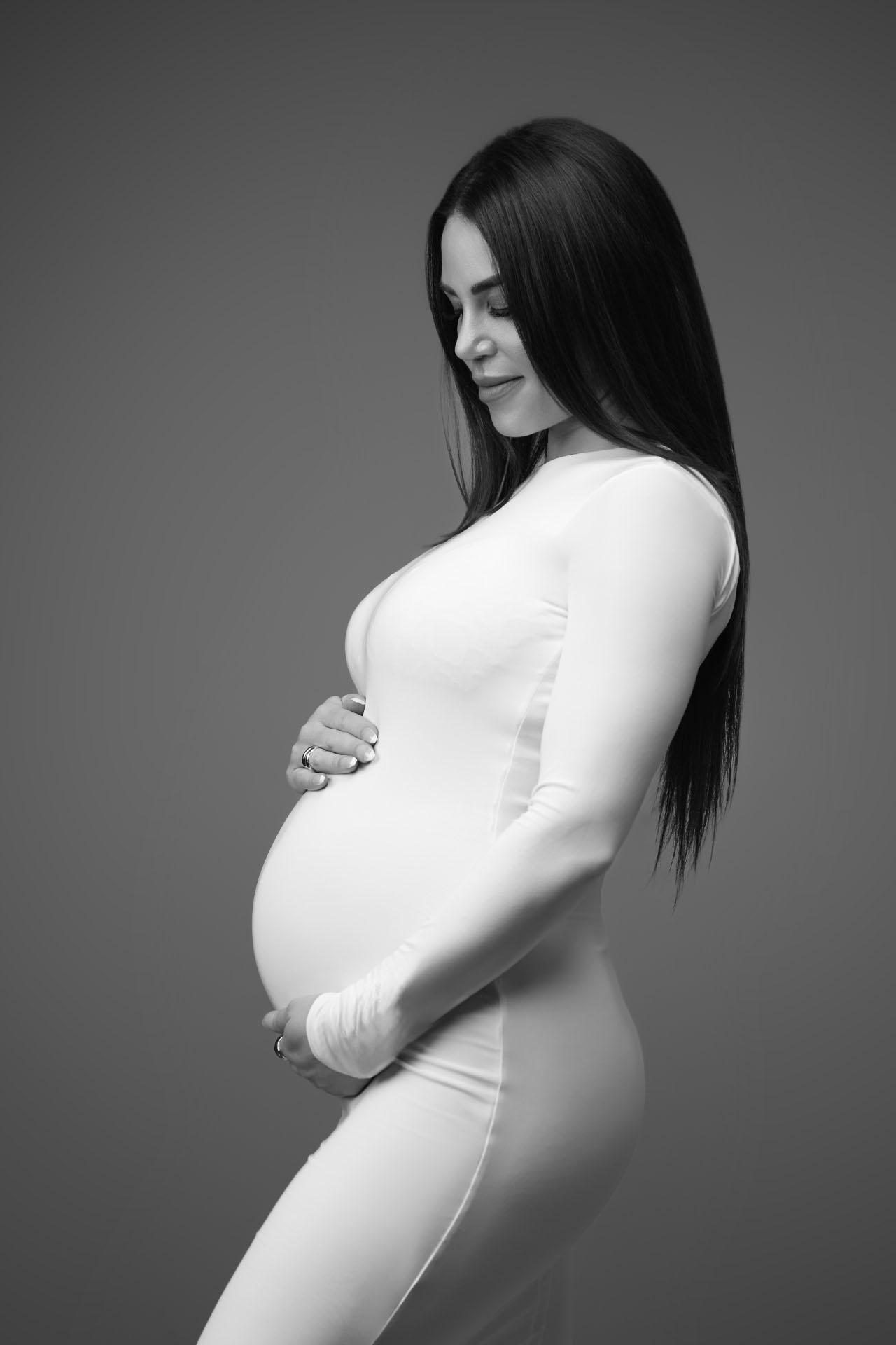 En gravid kvinde i en tætsiddende, langærmet hvid kjole står i profil, vugger forsigtigt sin mave og smiler blidt mod en ensfarvet baggrund - et fredfyldt barselsøjeblik, der er perfekt til et fotoshoot med Weigang Fotografi.