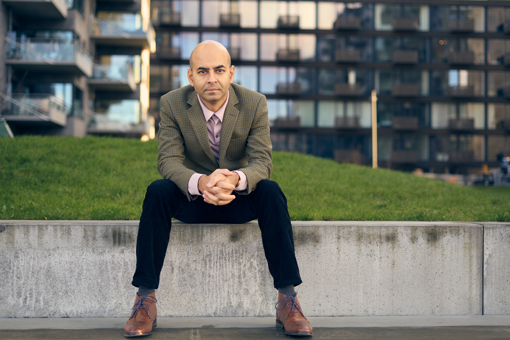 En mand i blazer og pæne sko sidder på en betonkant med græs bag sig, hænderne foldet, og kigger ind i kameraet. I baggrunden ses moderne boligblokke - en klassisk portrætforretning af Weigang Fotografi.