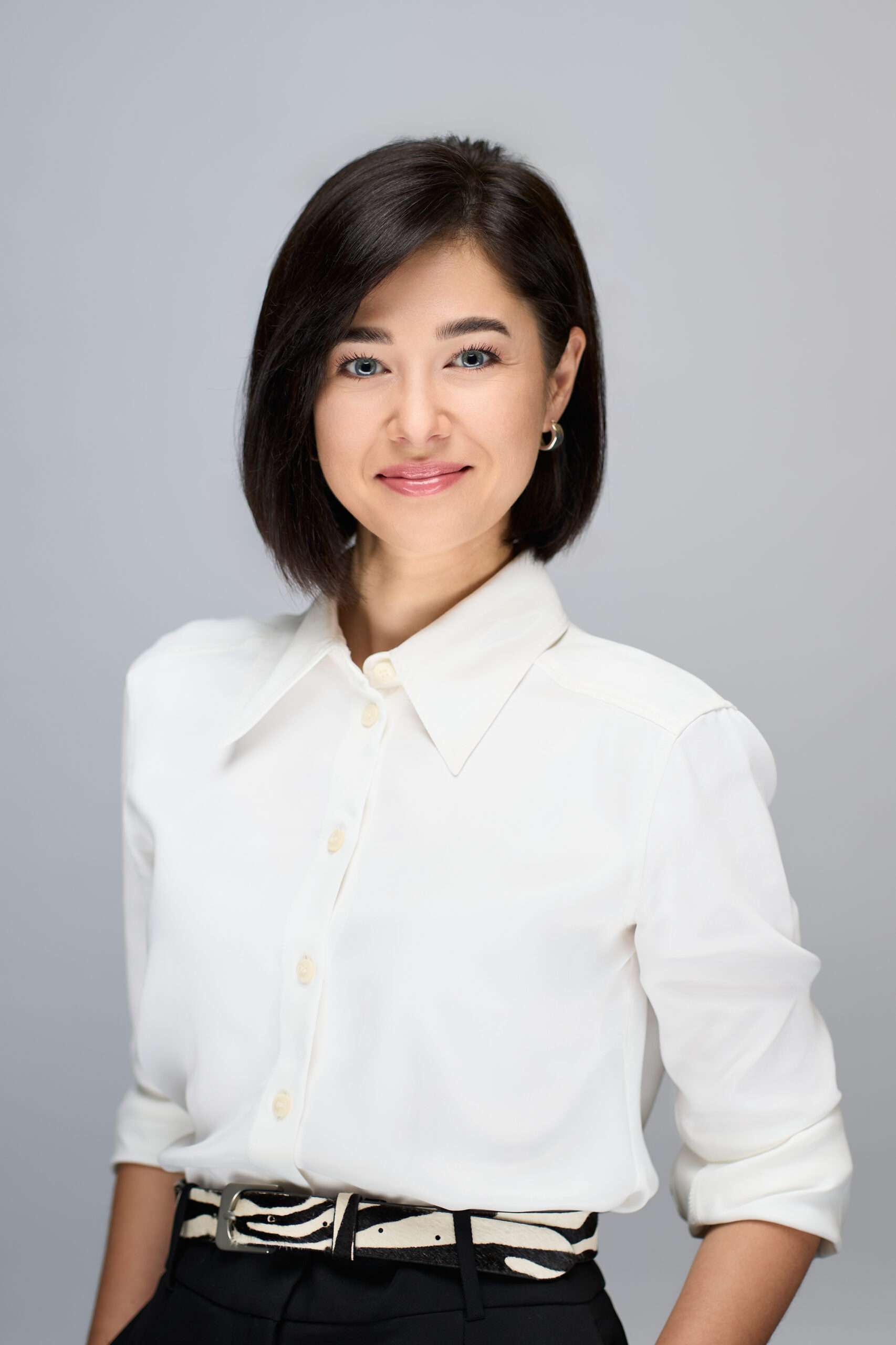 Kvinde med glat mørkt hår i hvid skjorte med knapper og sorte bukser, der smiler selvsikkert til sit Linkedin-billede på en almindelig lysegrå baggrund.