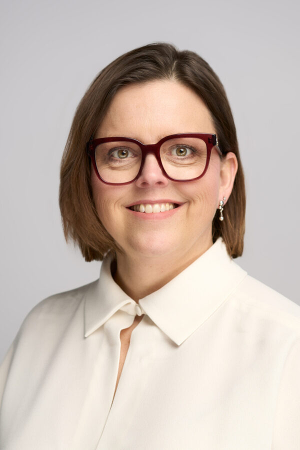 En kvinde med kort brunt hår, iført store røde briller, en hvid bluse med krave og øreringe, smiler til kameraet på en almindelig lysegrå baggrund.