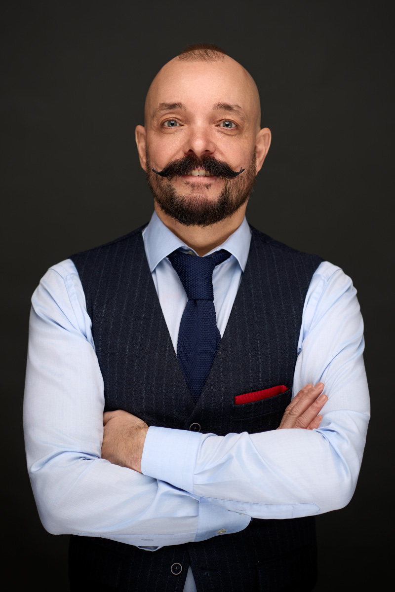 En skaldet mand med krøllet overskæg, iført lyseblå skjorte, marineblå vest og slips, står med armene over kors på en mørk baggrund. Han er perfekt til en Linkedin-portrætvirksomhed fra Weigang Fotografi og har en rød lommeklud og et selvsikkert udtryk.