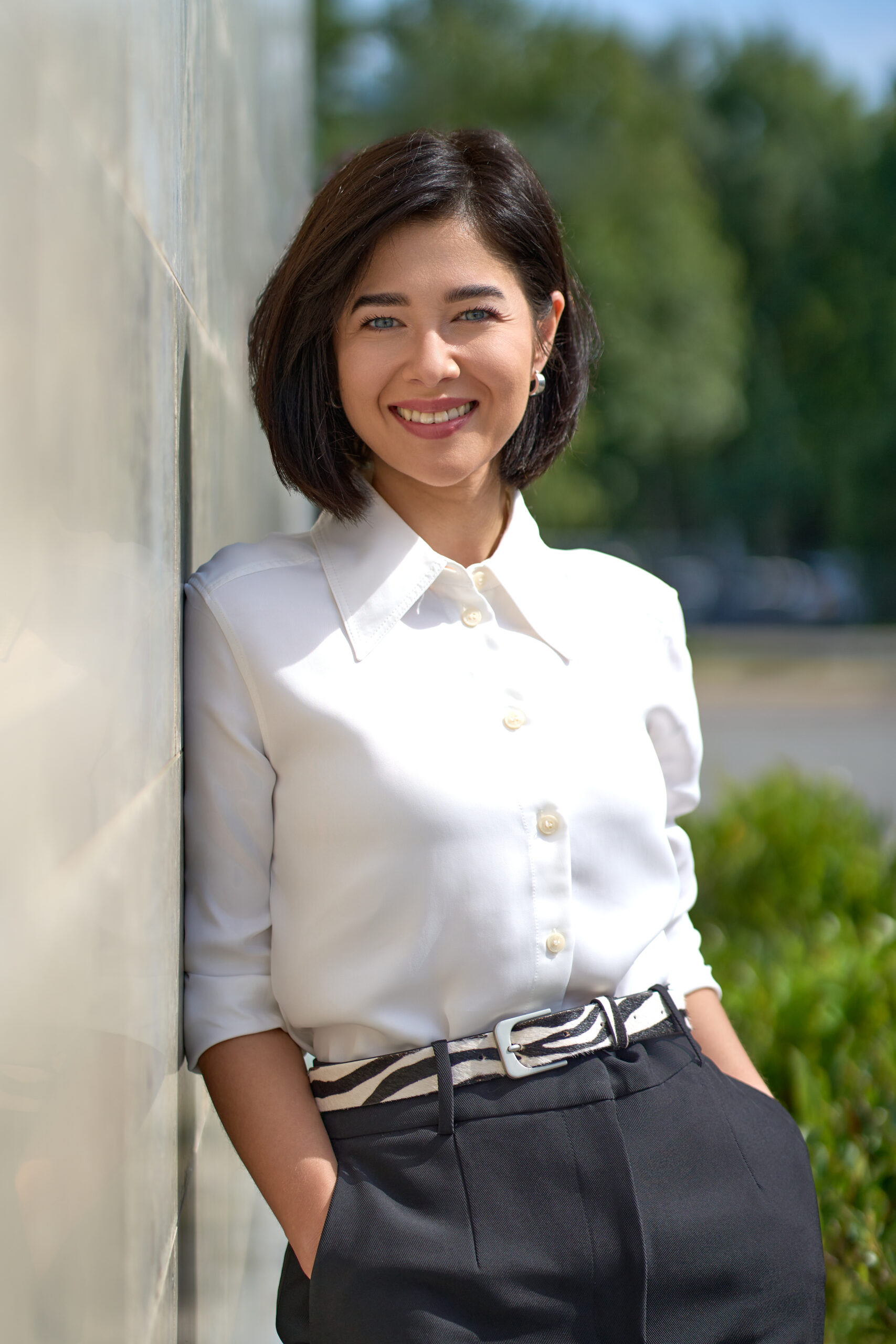En kvinde med kort mørkt hår, klædt i en hvid skjorte med knapper og sorte bukser, står udendørs og smiler og læner sig op ad en væg - et perfekt Linkedin-portræt til et forretningsmiljø med grønne planter og en sløret baggrund bag hende.